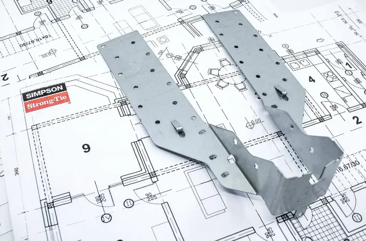 Metalowe łączniki Simpson Strong-Tie na tle planów architektonicznych. Pokazują, jak łączyć drewno konstrukcyjne, zapewniając stabilność i bezpieczeństwo budowy.