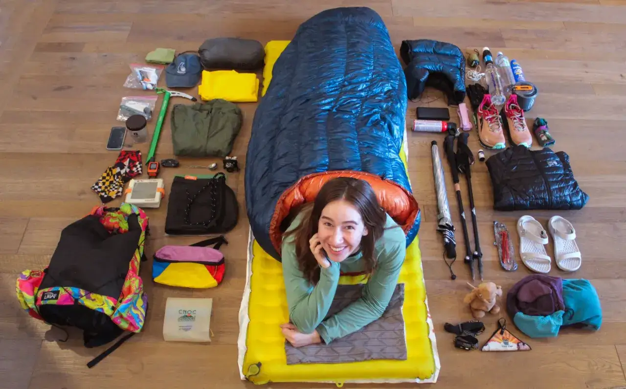 Uśmiechnięta dziewczyna leży w śpiworze puchowym czy syntetycznym, otoczona ekwipunkiem turystycznym: plecak, buty, kijki, odzież.
