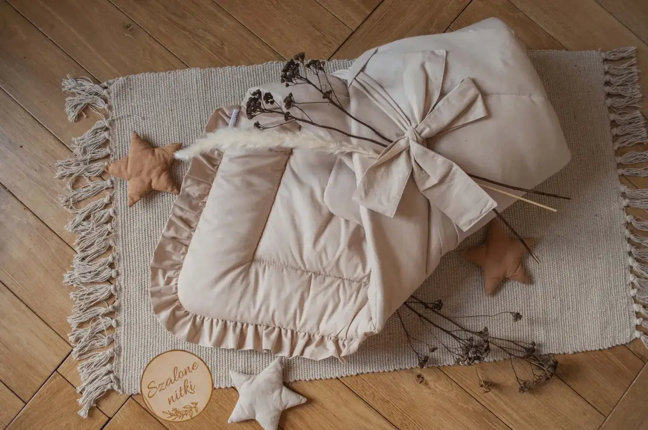 Beżowy rożek niemowlęcy z falbanką, ozdobiony kokardą i suszonymi gałązkami, leży na dywaniku z frędzlami.