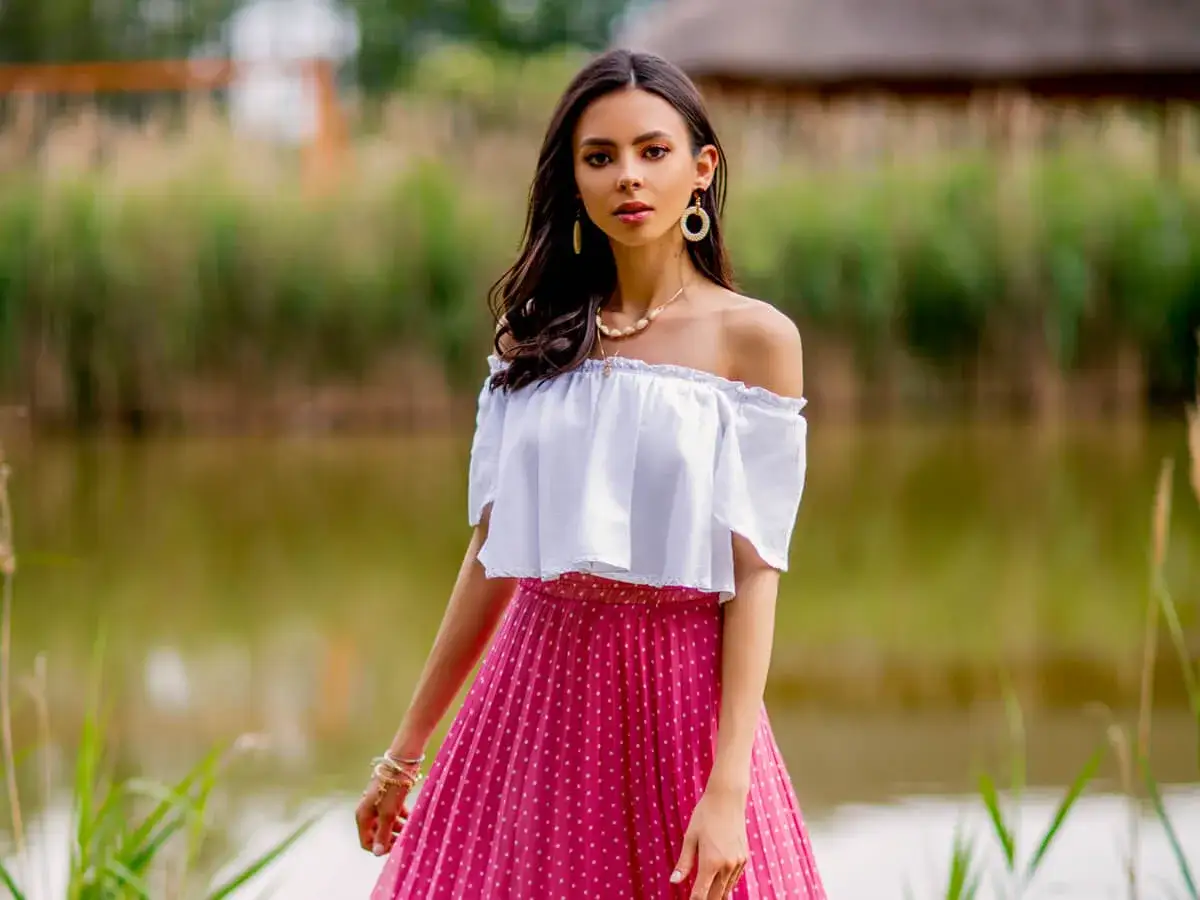Kobieta w białej bluzce hiszpance na lato i różowej spódnicy w groszki pozuje nad wodą.