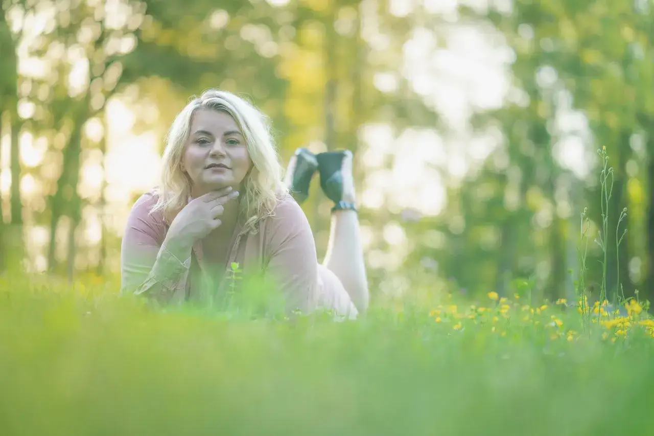Kobieta w r&oacute;żowej sukience leży na trawie podczas sesji biznesowej w plenerze. W tle drzewa i ż&oacute;łte kwiaty.