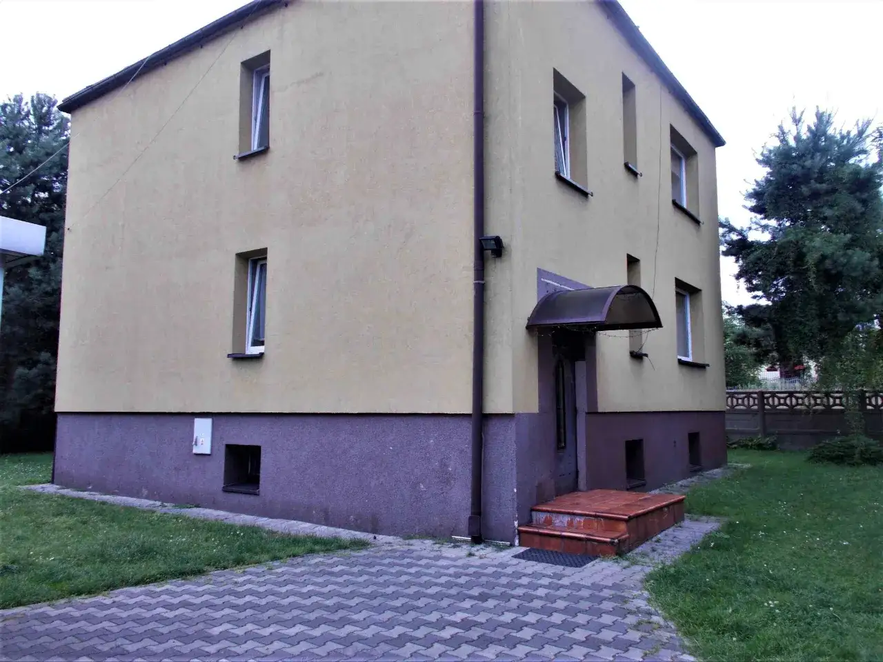 Domy wystawione na licytacje przez komornika. Budynek z ż&oacute;łtym tynkiem i brązowym cokołem, z kilkoma oknami i wejściem pod daszkiem.