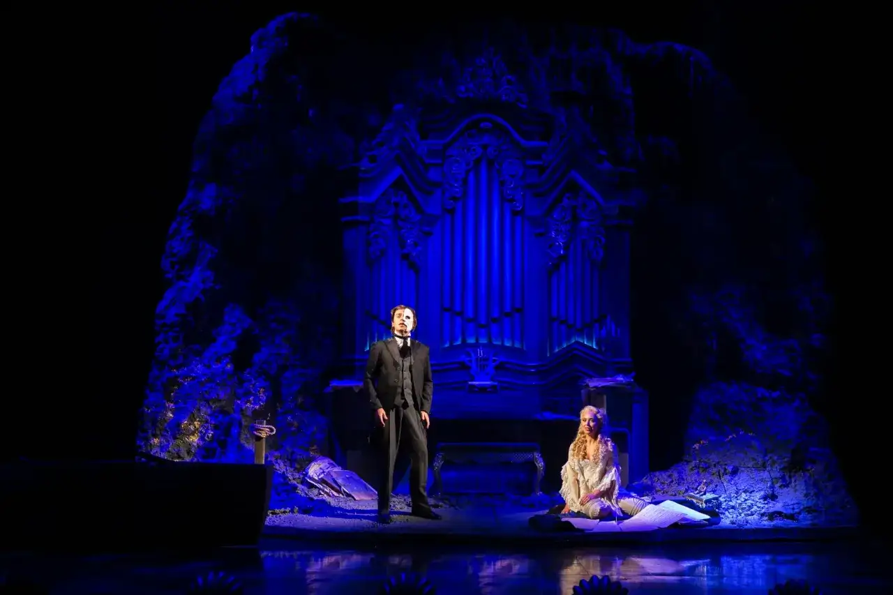 Upiór w operze w Warszawie. Tajemnicza postać w masce stoi obok siedzącej Christine na tle niebieskich organów.