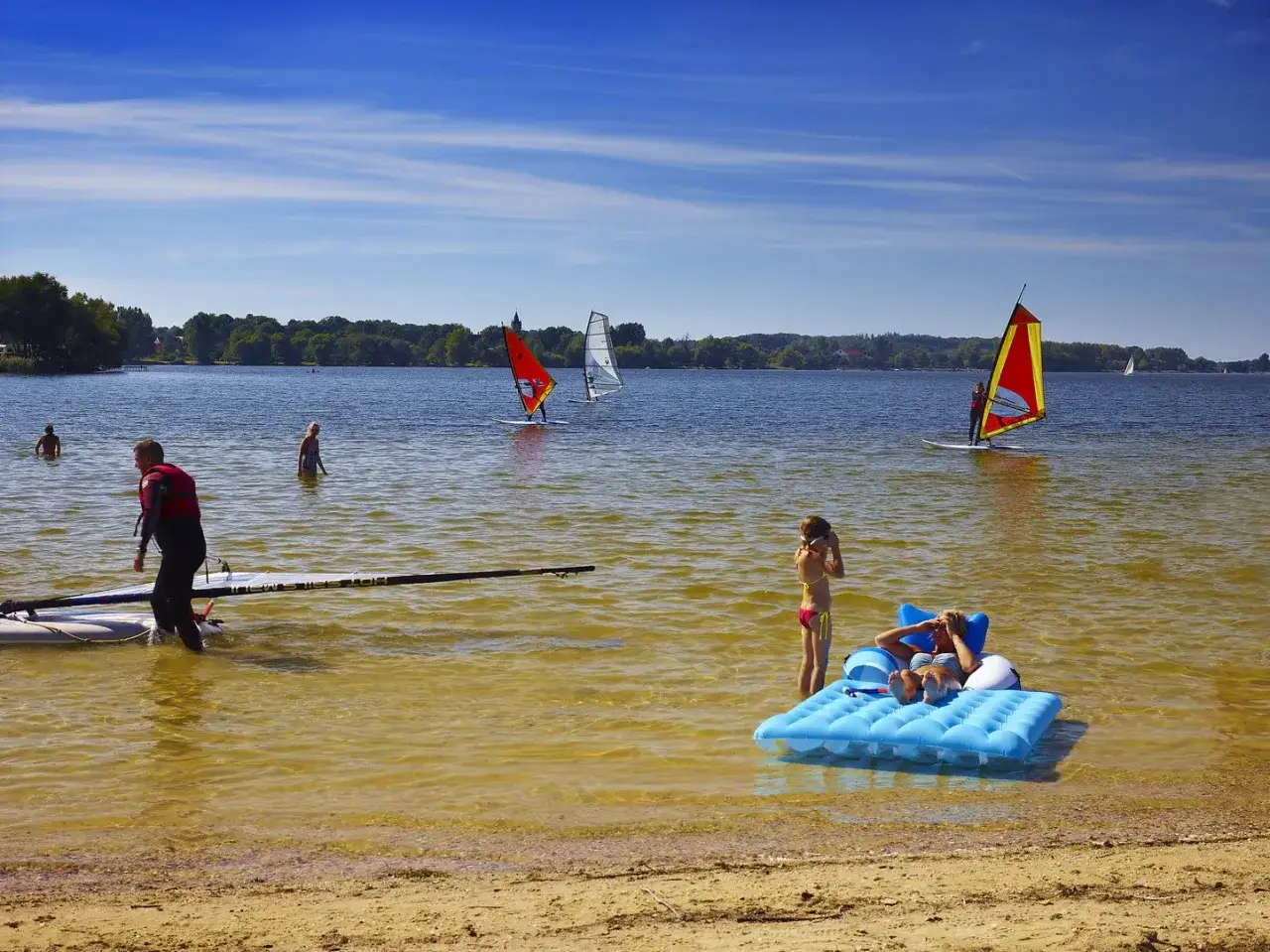 Na jeziorze Pławniowice wiatr napędza żagle desek windsurfingowych. Na brzegu odpoczywają ludzie.