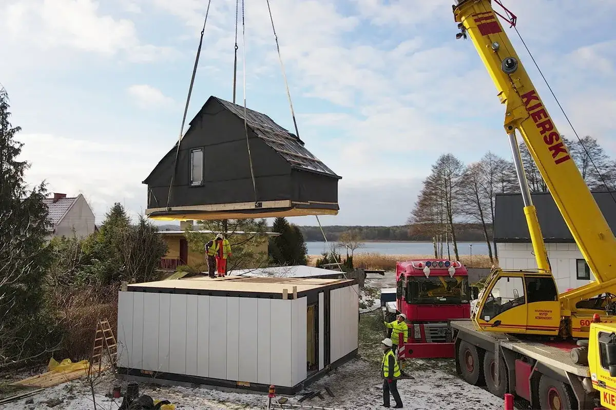 Montaż dom&oacute;w modułowych bez pozwolenia. Dźwig unosi gotowy moduł dachu na budynek.