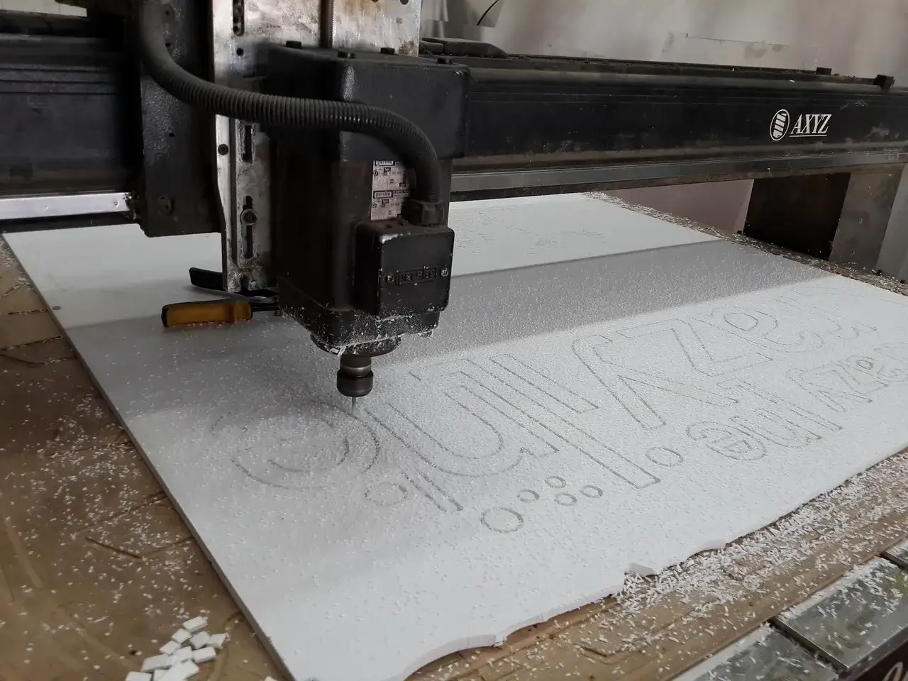 Ploter CNC DIY wycina napisy na białej płycie. Wi&oacute;ry tworzą biały dywan wok&oacute;ł frezu.