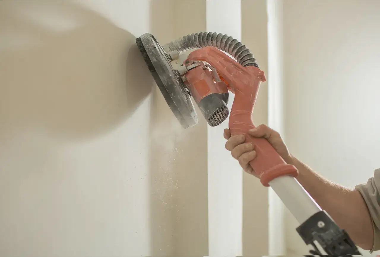 Ręka trzyma szlifierkę do ścian, która podczas pracy generuje pył. To pokazuje, jak nakładać gładź szpachlową, by uzyskać idealnie gładką powierzchnię.