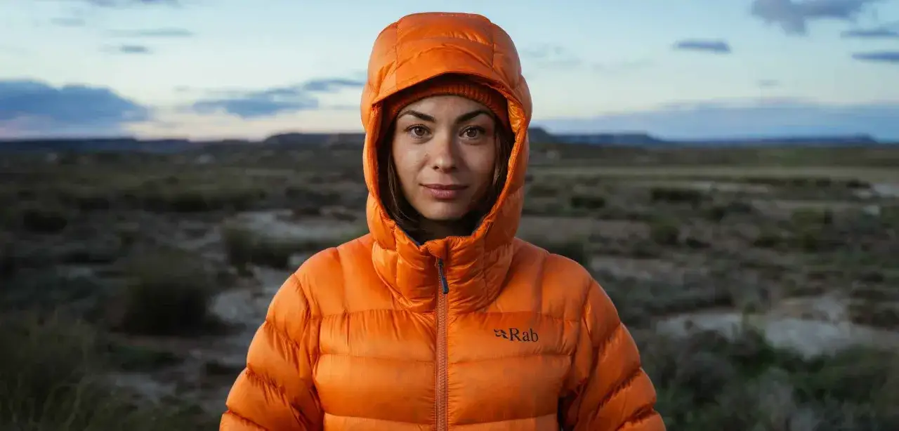 Kobieta w pomarańczowej, puchowej kurtce Rab, idealnej jako jaka kurtka na zimę, na tle pustynnego krajobrazu.