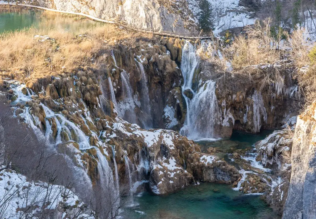 Zimowe Jeziora Plitwickie: wodospady spływają po skalnych tarasach, a l&oacute;d i śnieg tworzą lodowe sople. Drewniana kładka wije się nad turkusową wodą.