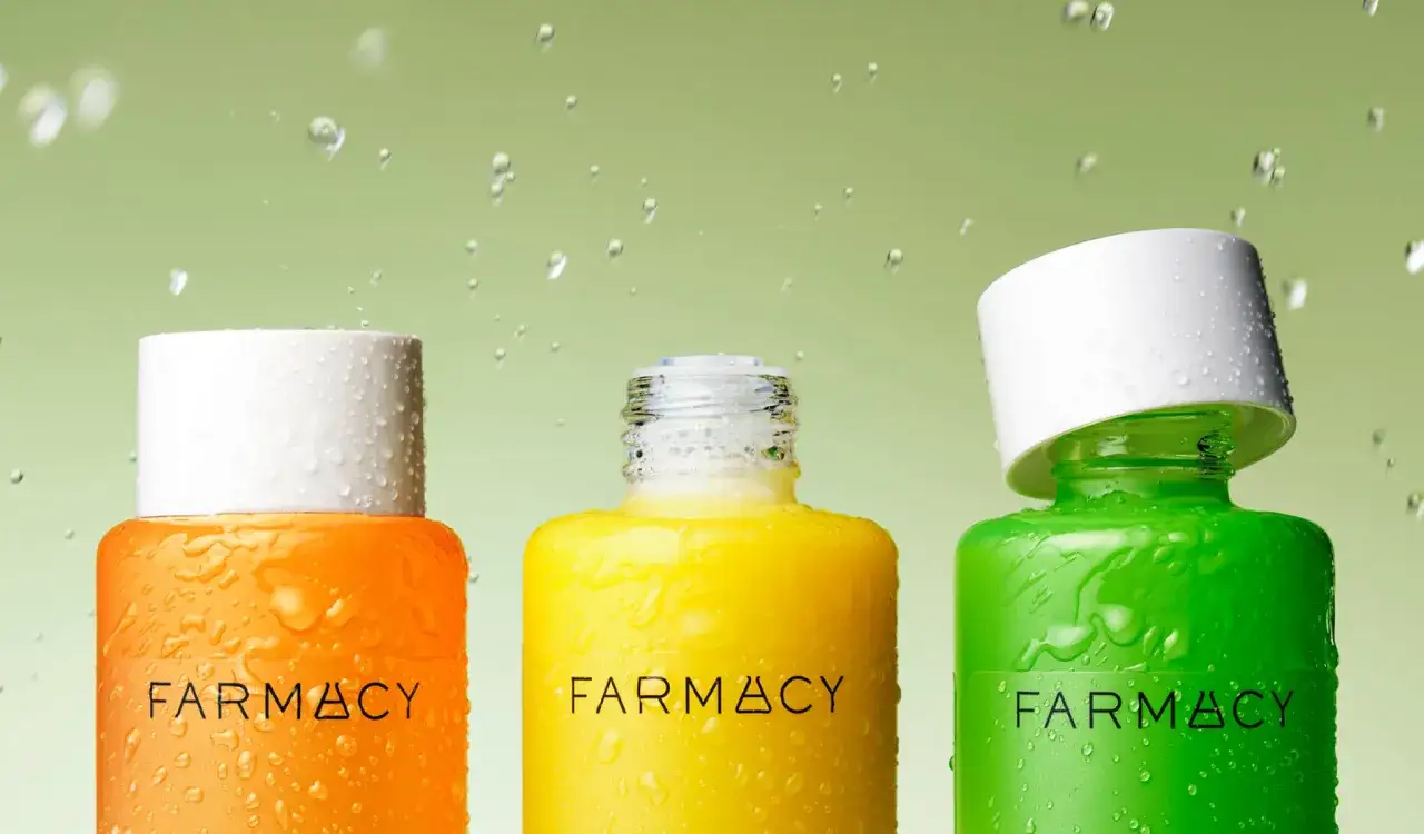 Trzy kolorowe butelki Farmacy: pomarańczowa, ż&oacute;łta i zielona, pokryte kroplami wody. Każda zawiera esencję do twarzy, gotową do odświeżenia sk&oacute;ry.