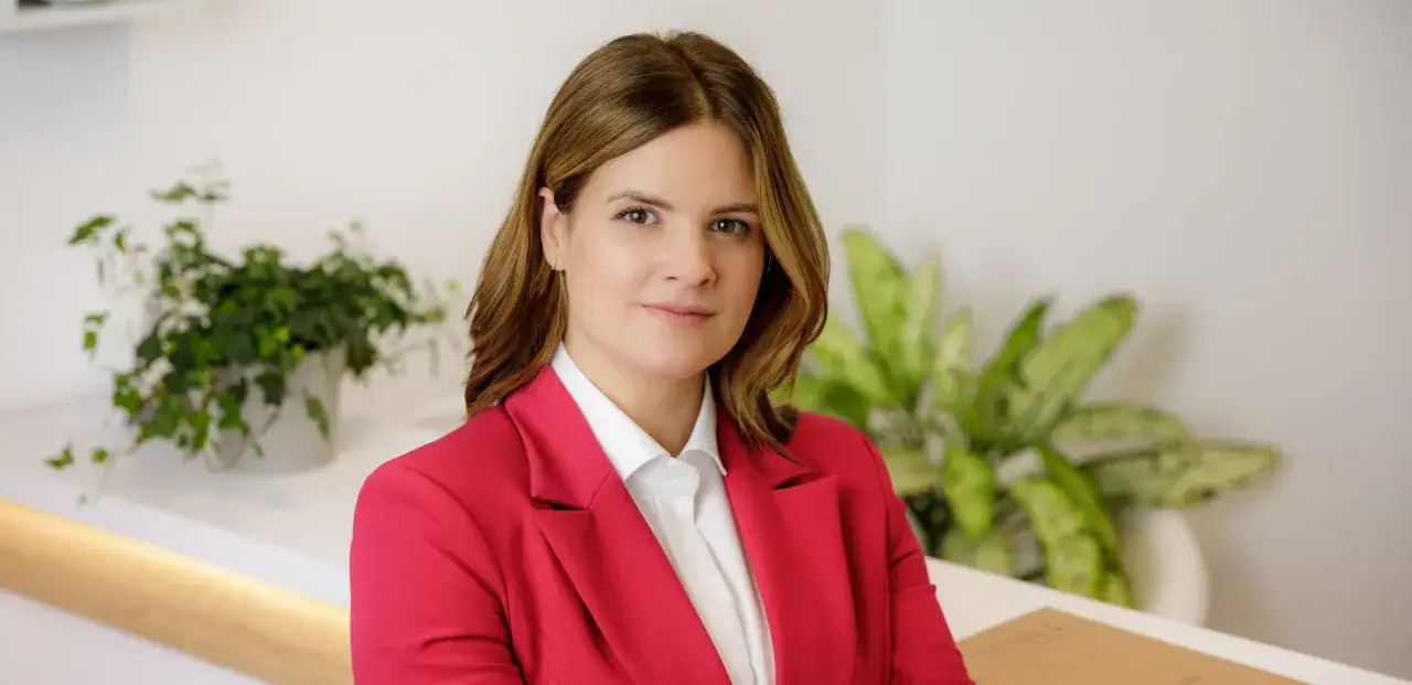 Kobieta w czerwonej marynarce i białej koszuli, w tle rośliny. Może to być notariusz przygotowujący testament notarialny.