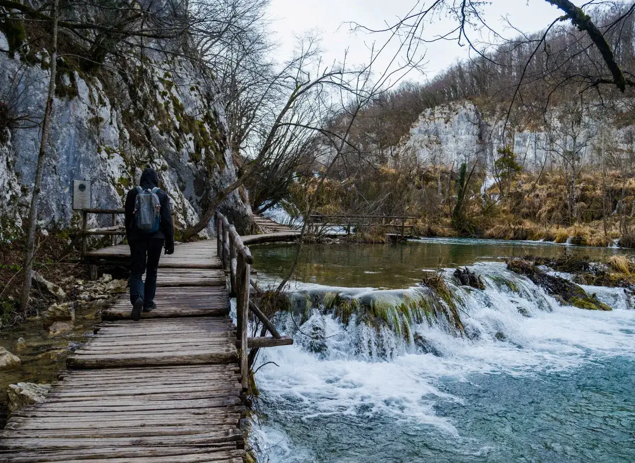 Spacer przemierza drewniane kładki nad wodospadami. Jeziora Plitwickie zimą, w otoczeniu nagich drzew i skalnych ścian, tworzą surowy, piękny krajobraz.