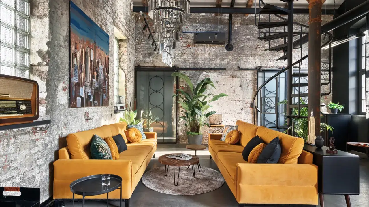 Wnętrze w **stylu loftowym w bloku** z ceglanymi ścianami, ż&oacute;łtymi sofami, spiralnymi schodami i dużą rośliną.