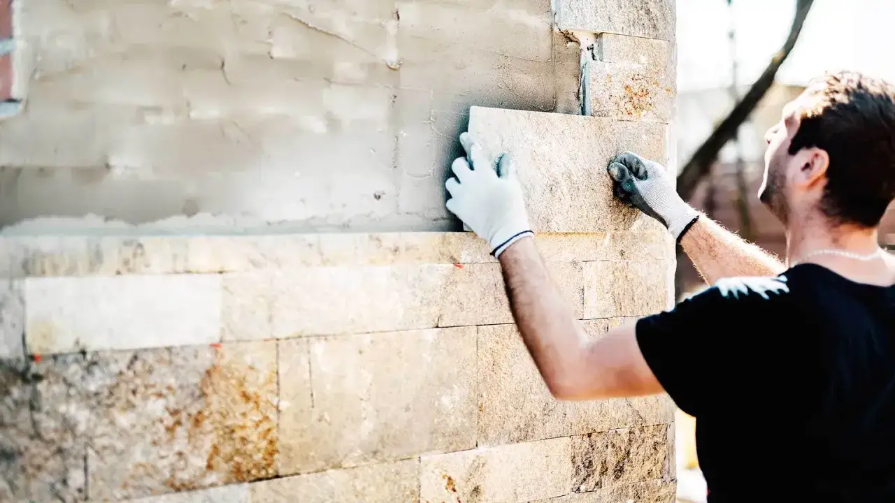 Pracownik w rękawiczkach układa kamienne płytki, pokazując, jak odnowić elewację domu.