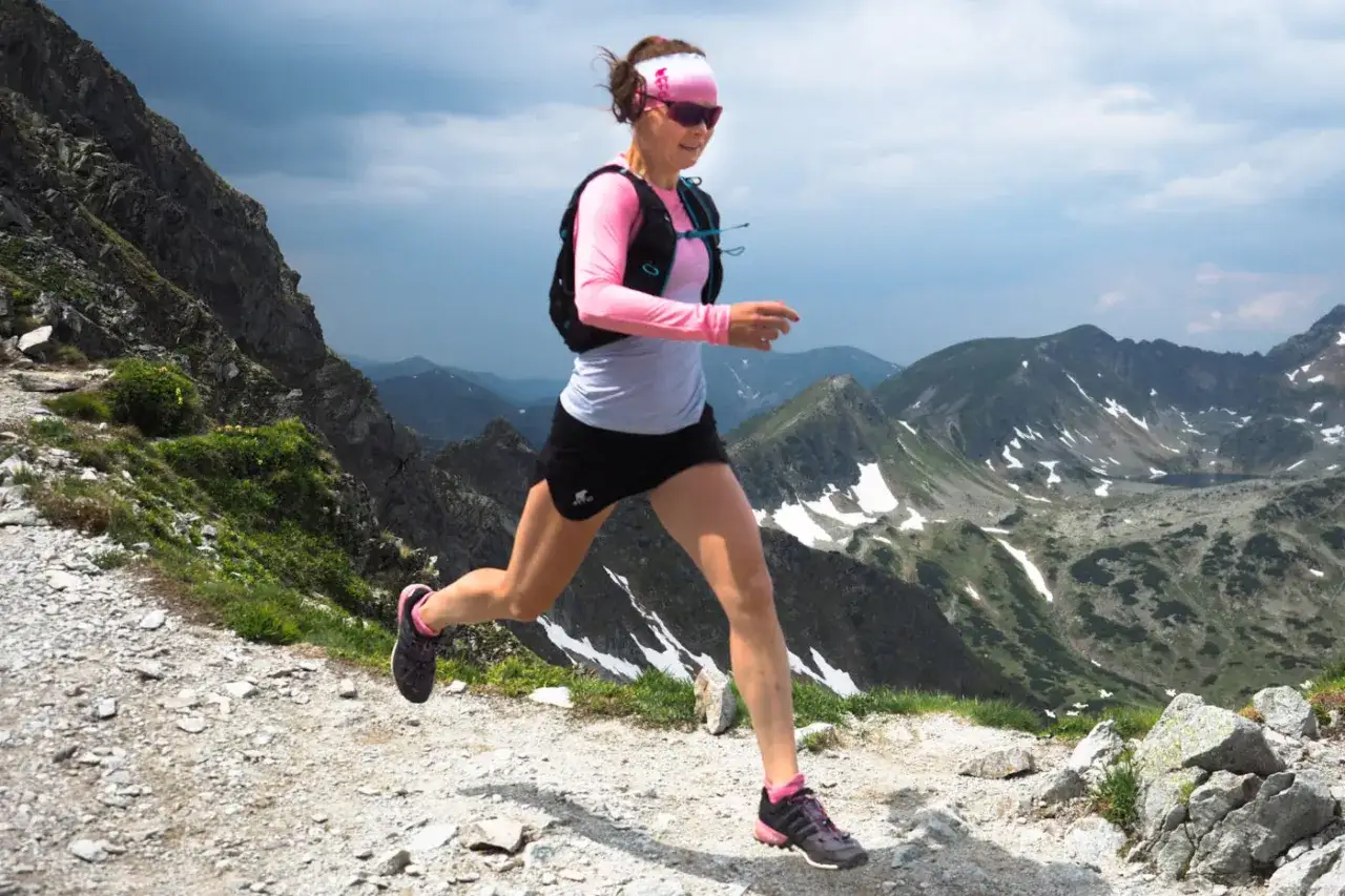 Kobieta biegnie po g&oacute;rskim szlaku, używając kij&oacute;w do biegania po g&oacute;rach. W tle piękne, skaliste krajobrazy.