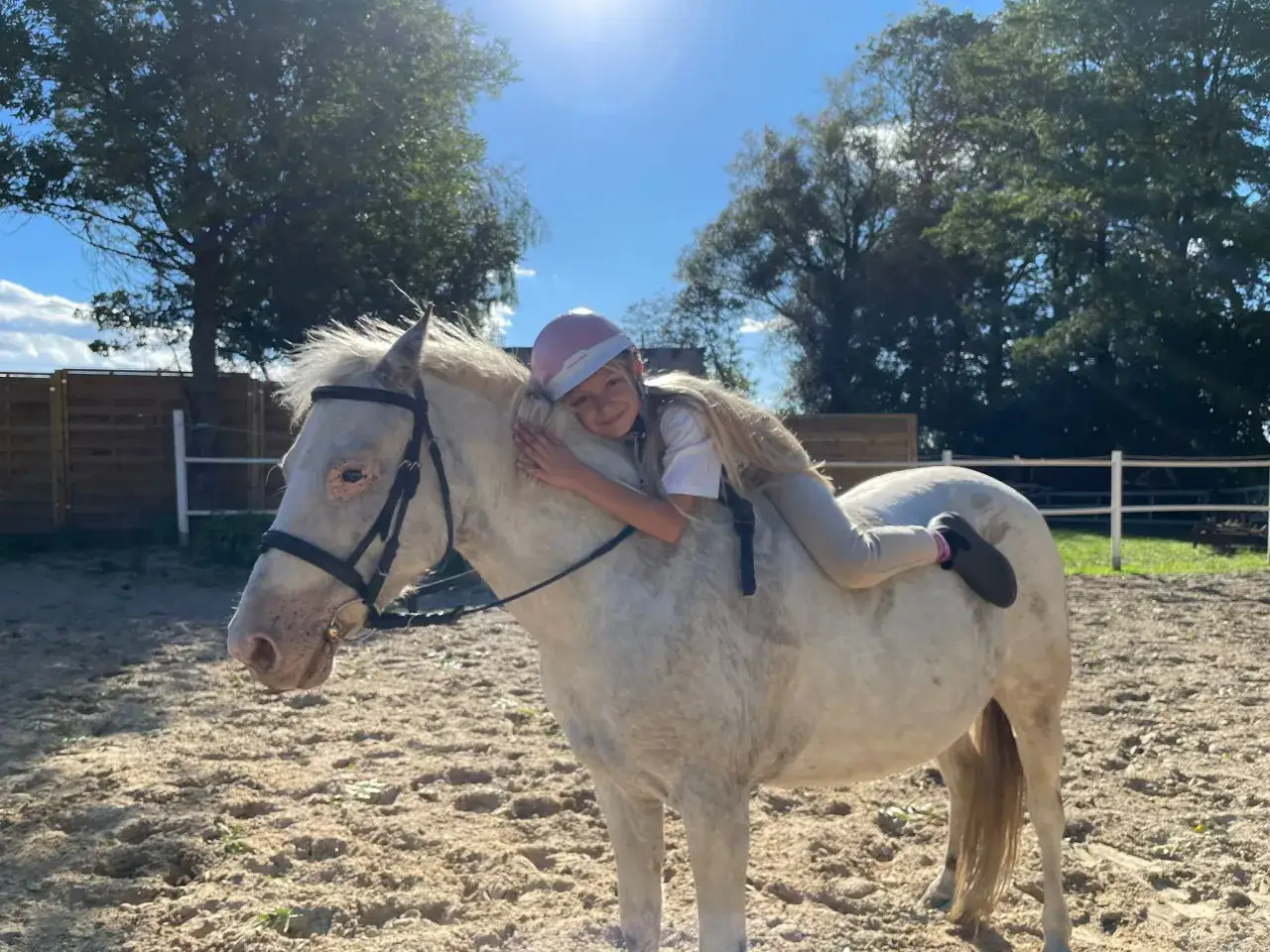 Dziewczynka przytula się do siwego konia na ujeżdżalni. To ich wspólna przygoda w stajni, gdzie każdy koń to ostoja spokoju, a każda kończyna to krok do wolności.
