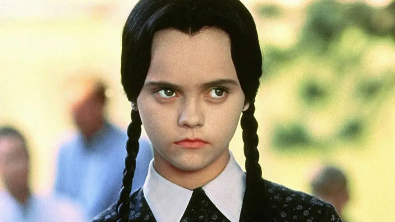 Christina Ricci jako Wednesday Addams, z charakterystycznymi warkoczami i ponurym wyrazem twarzy, wpatruje się w dal.