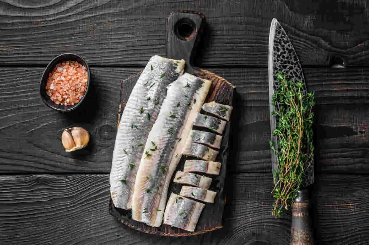 Filety śledziowe na desce, obok n&oacute;ż z ziołami, czosnek i s&oacute;l. Czy pies może jeść śledzie?