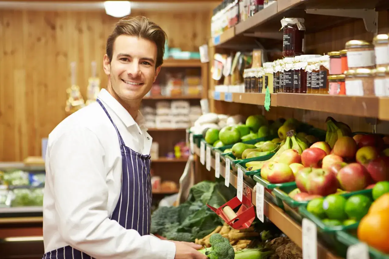 Uśmiechnięty właściciel małego sklepu spożywczego, otoczony świeżymi owocami i warzywami, zastanawia się, ile zarabia po odliczeniu podatków.