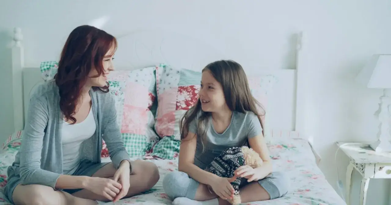 Mama rozmawia z c&oacute;rką na ł&oacute;żku. Dziewczynka trzyma pluszowego misia. To może być rozmowa o pierwszym telefonie dla dziecka.