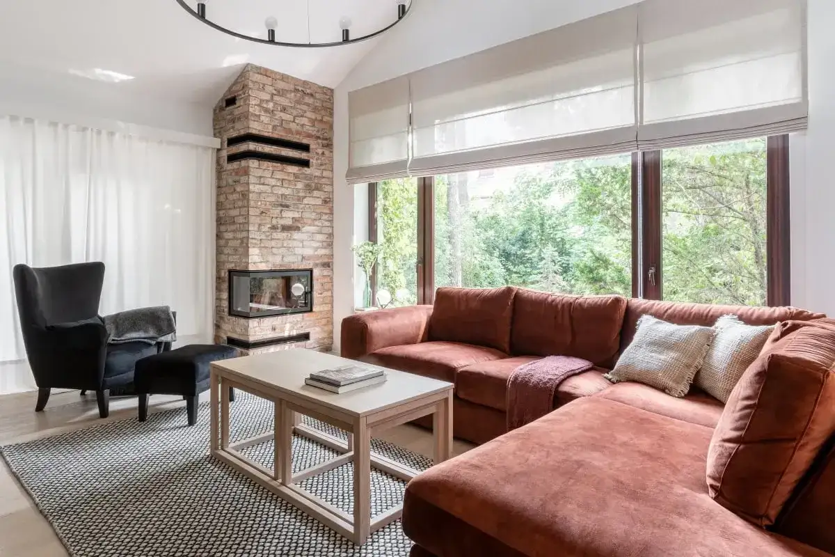 Przytulny salon w stylu holenderskim z ceglanym kominkiem, dużym oknem i wygodną, rudą kanapą.