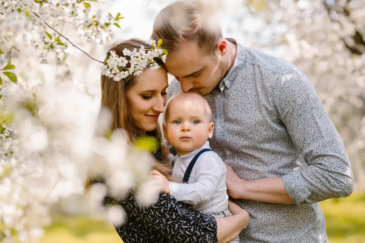 Uśmiechnięta rodzina w kwitnącym sadzie. Mama z wiankiem z kwiat&oacute;w, tata całuje dziecko w czoło. To była śmieszna sesja rodzinna.