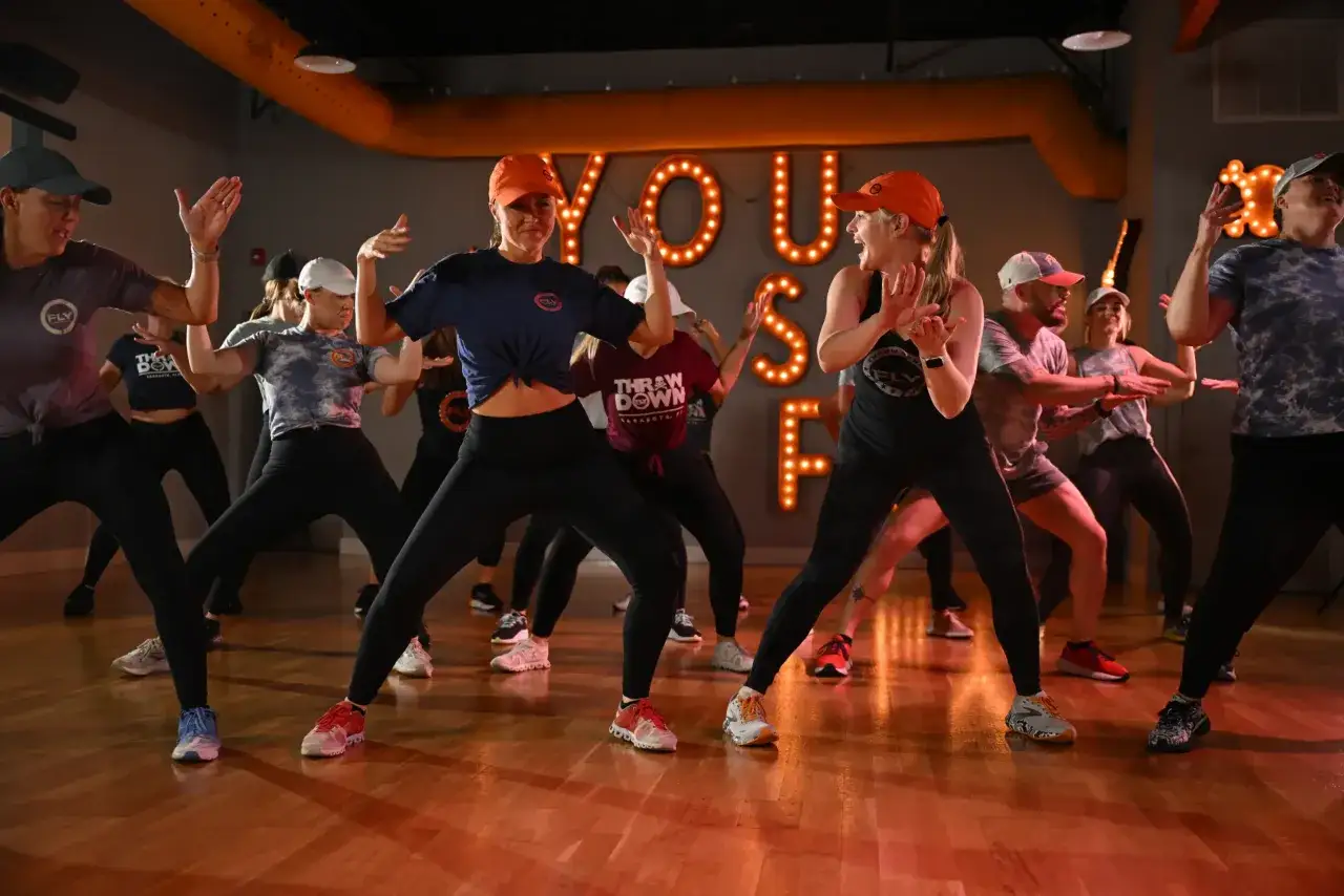 Grupa os&oacute;b ćwiczy podstawowe kroki zumby w sali z podświetlanym napisem.