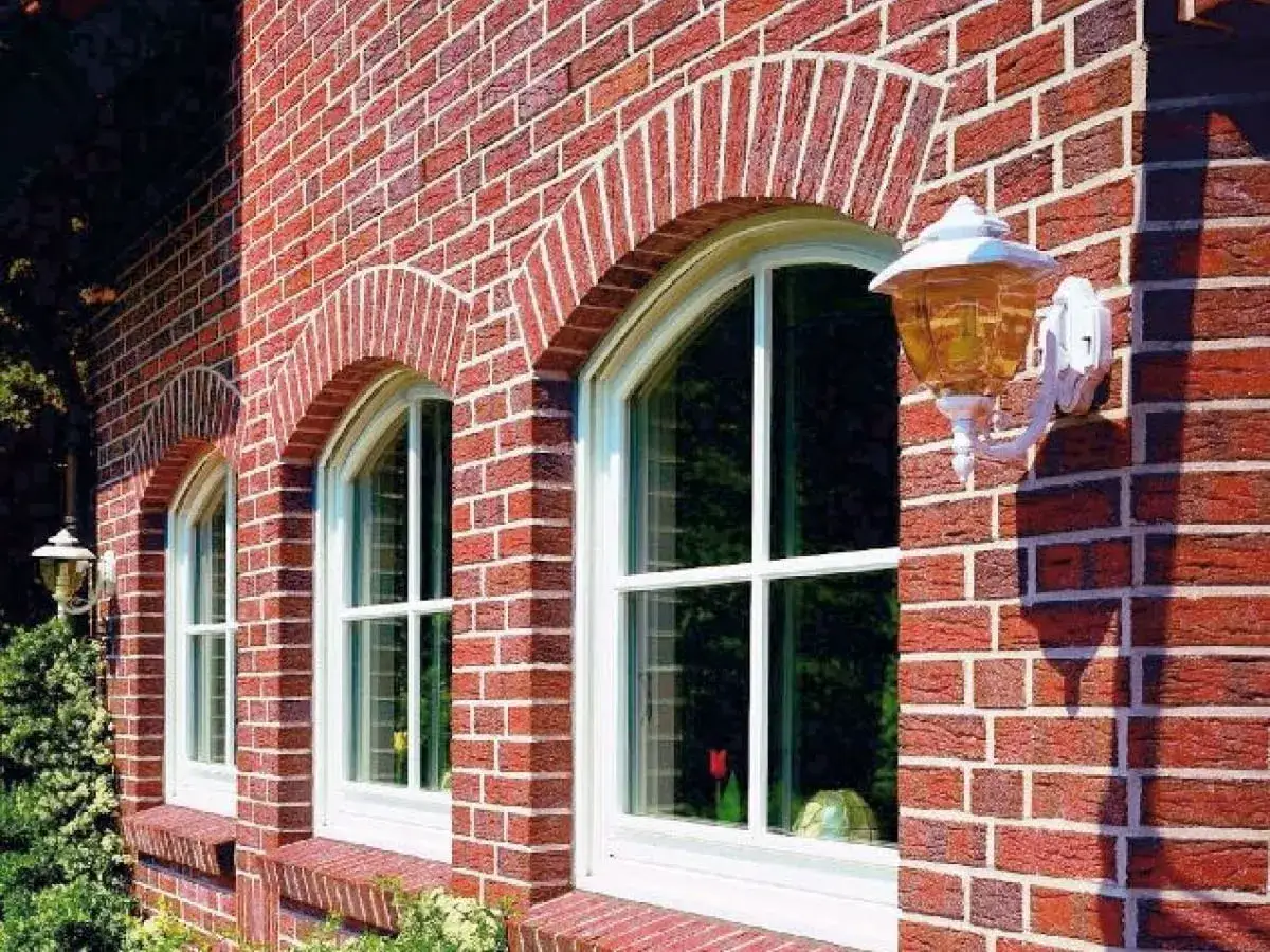 Elegancki łuk nad oknem zdobi czerwoną ceglaną ścianę. Obok wisi stylowa lampa.