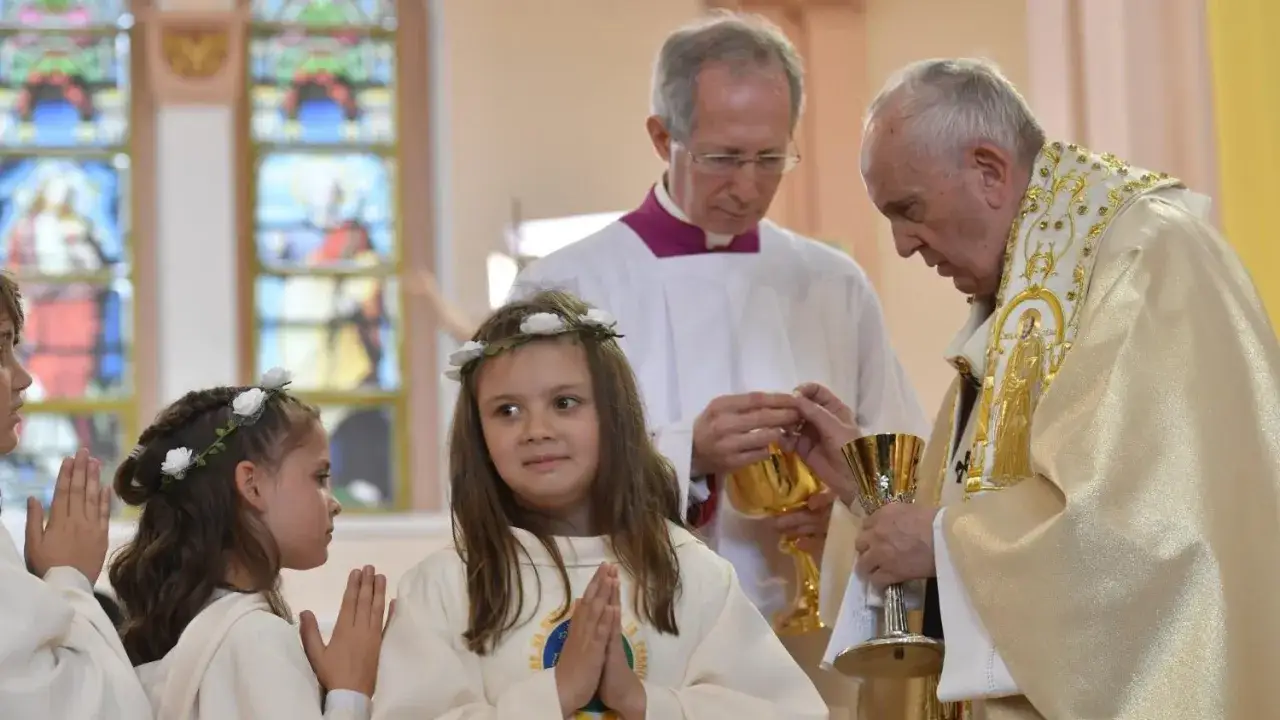 Dzieci w białych szatkach, z wiankami na głowach, modlą się podczas pierwszej modlitwy eucharystycznej. Papież Franciszek udziela Komunii.