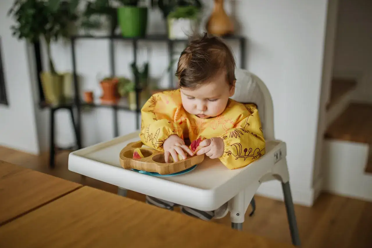 Niemowlę w ż&oacute;łtym śliniaku pr&oacute;buje sięgnąć po pierwsze owoce dla niemowlaka z drewnianej miseczki.