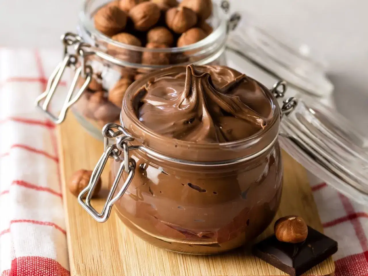 Słoik z kremem ferrero rocher, obok orzechy laskowe i kawałek czekolady.