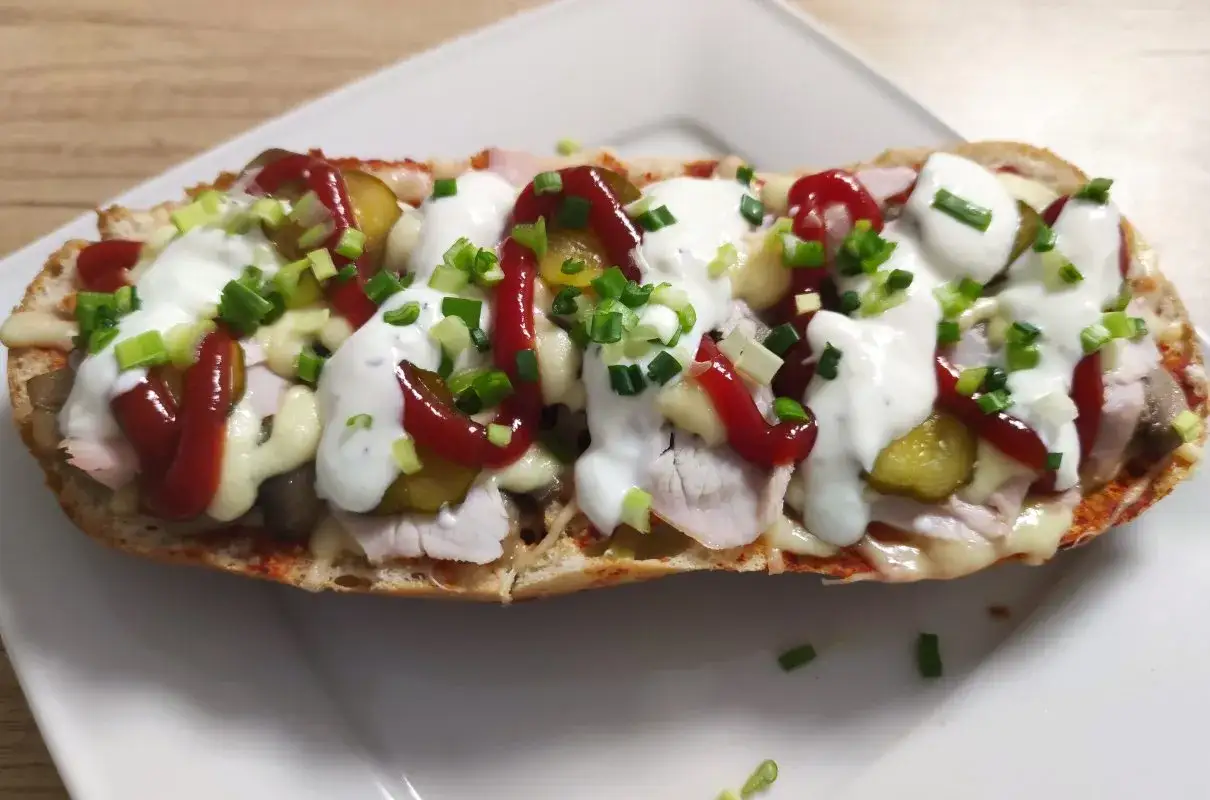 Pyszna zapiekanka z bagietki z serem, szynką, ogórkiem, keczupem i majonezem, posypana szczypiorkiem.