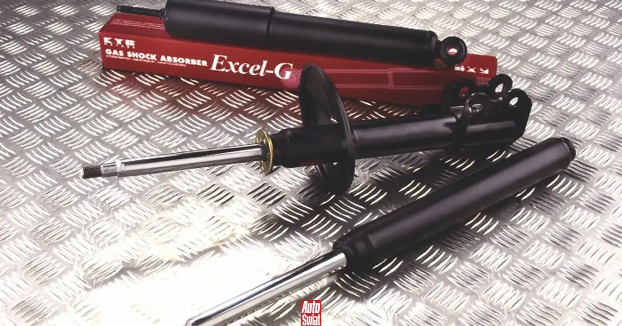 Różne rodzaje amortyzatorów samochodowych, w tym amortyzator KYB Excel-G, leżą na metalowej powierzchni.