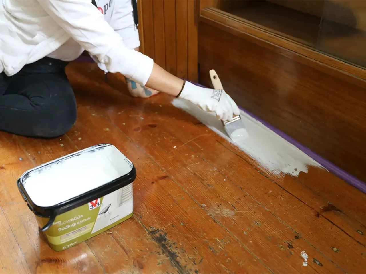Osoba w białej rękawiczce maluje pędzlem starą podłogę drewnianą na jasny kolor. Obok stoi wiaderko z farbą.