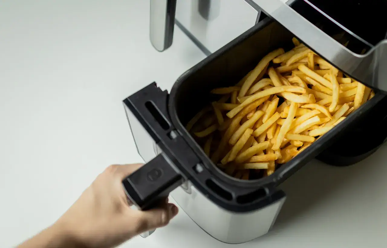 Ręka wyciąga koszyk z chrupiącymi frytkami z frytkownicy beztłuszczowej, sugerując, czy pieczone frytki są zdrowe.