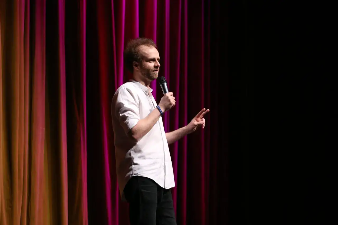 Marcin Zbigniew Wojciech stand up na scenie z mikrofonem, gestykulując do publiczności.