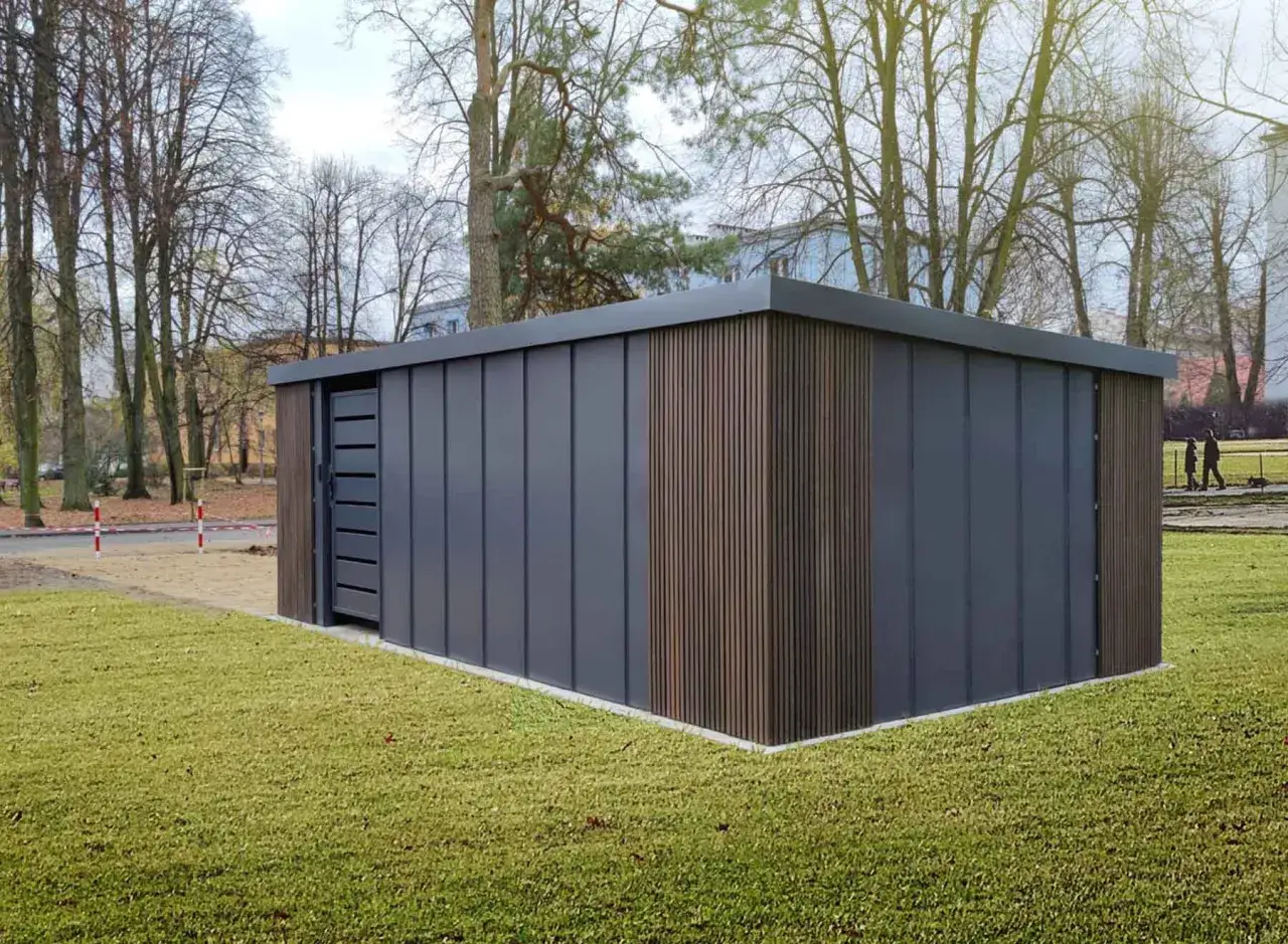 Nowoczesne miejsce na kosze na śmieci w ogrodzeniu, z drzwiami i panelami imitującymi drewno, otoczone zielonym trawnikiem i drzewami.