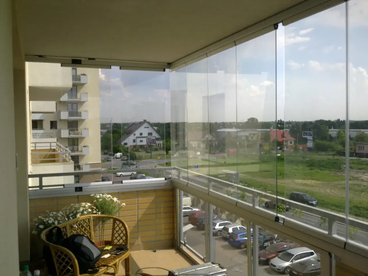 Zabudowany balkon w bloku ze szklanymi panelami, widok na osiedle i ulicę. Krzesło z poduszką i plecakiem.