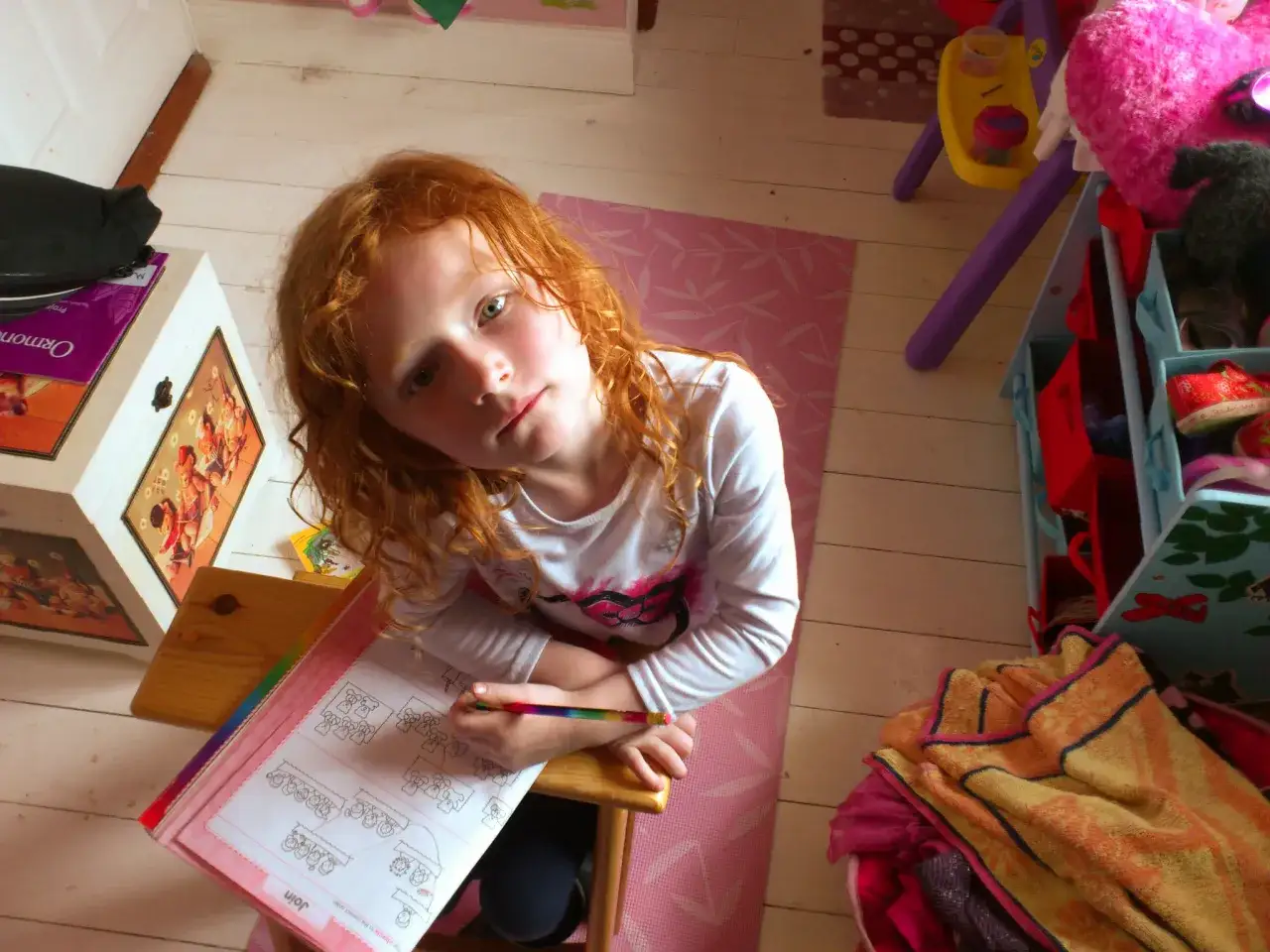 Ruda dziewczynka z zielonymi oczami z zapałem rozwiązuje zadania w książeczce, pokazując, jak zachęcić dziecko do nauki przez zabawę.