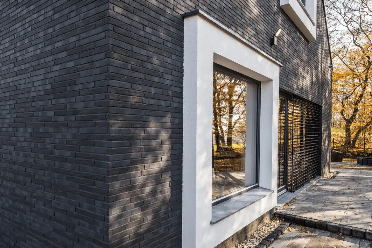 Nowoczesna elewacja z klinkieru w ciemnym kolorze, z białym obramowaniem okna i jesiennym krajobrazem w tle.