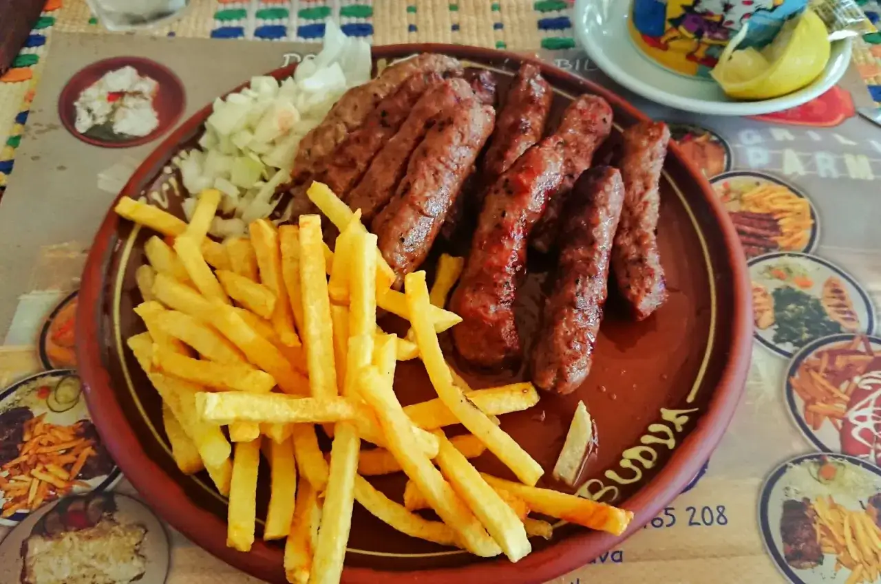 Czarnog&oacute;ra jedzenie: grillowane kiełbaski ćevapi z frytkami i posiekaną cebulą na brązowym talerzu.