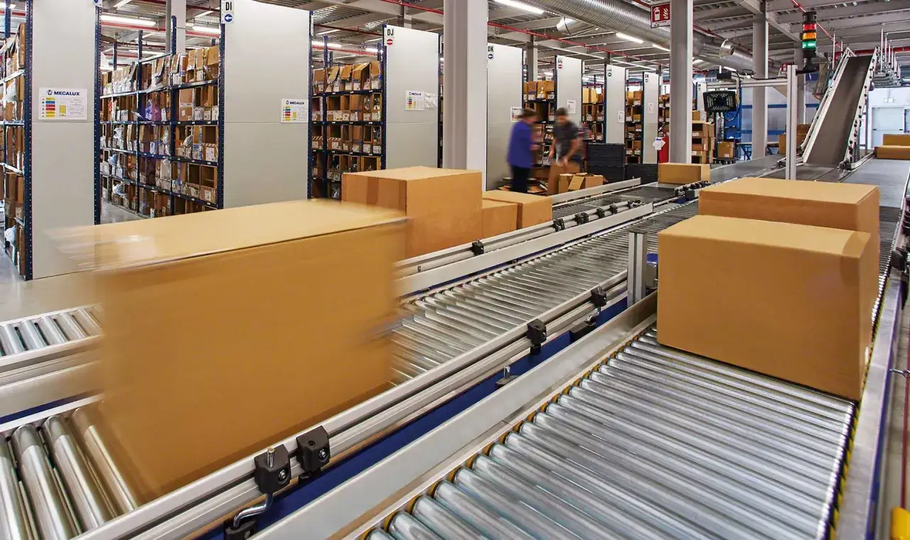 Magazyn z taśmociągami i kartonami. Widać regały z towarami, pracownik&oacute;w i ruchome paczki, kt&oacute;re czekają na realizację zam&oacute;wienia (back order).