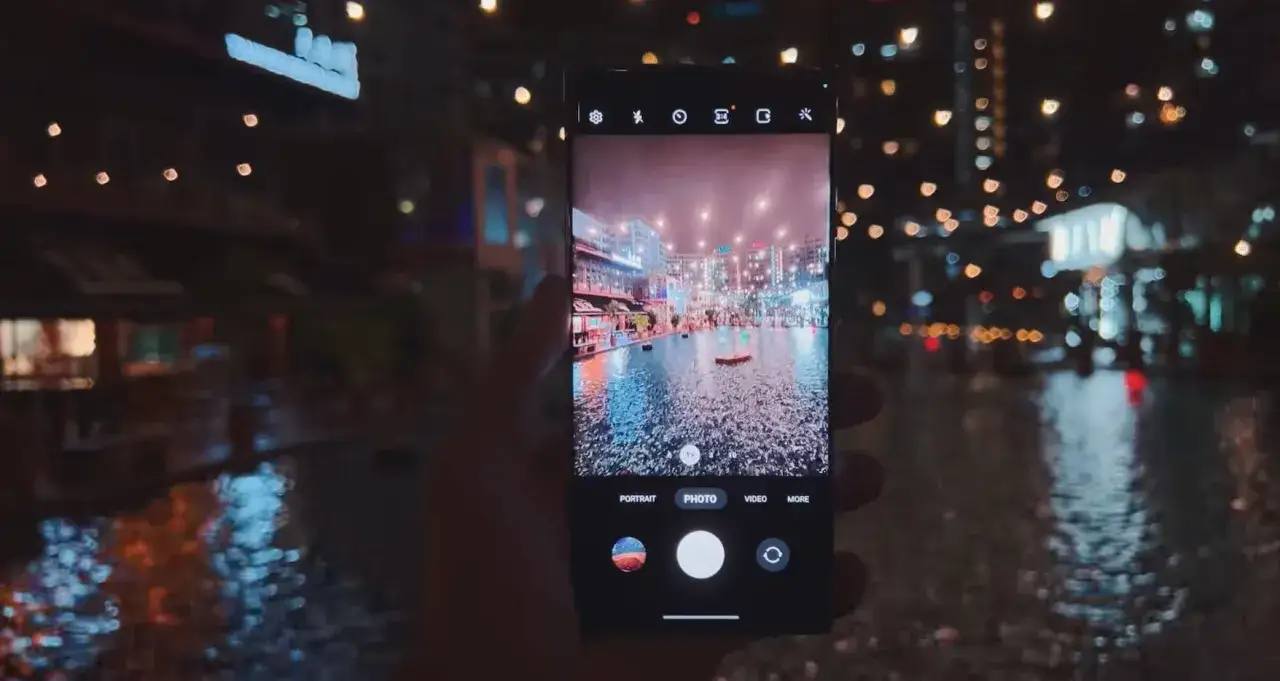 Ręka trzyma aparat cyfrowy Samsung Galaxy, fotografując nocny widok miasta z odbiciami świateł w wodzie.