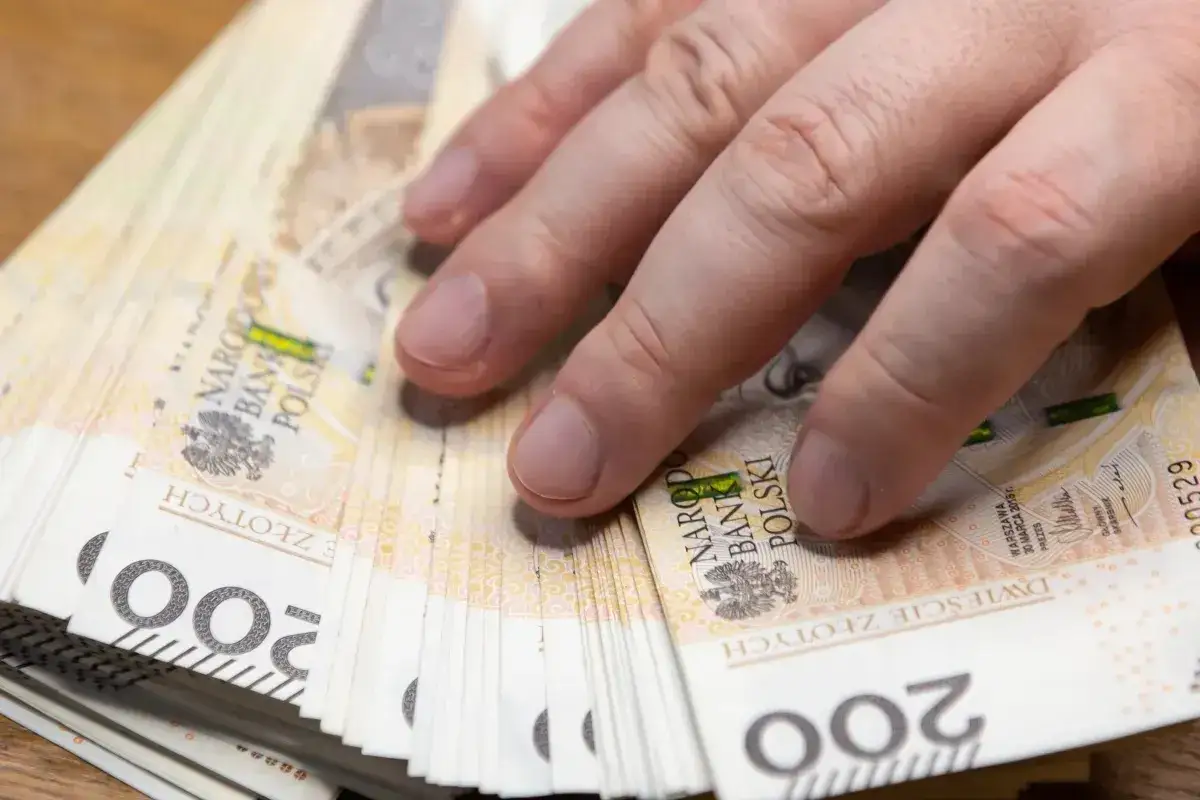 Dłoń na stosie banknotów 200 zł. Czy emeryta obowiązuje okres wypowiedzenia? Pieniądze to ważna sprawa.