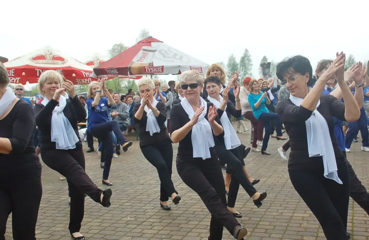 Radosna potańcówka dla seniorów w Warszawie. Kobiety w czerni i białych szalach tańczą na zewnątrz, pod parasolami.