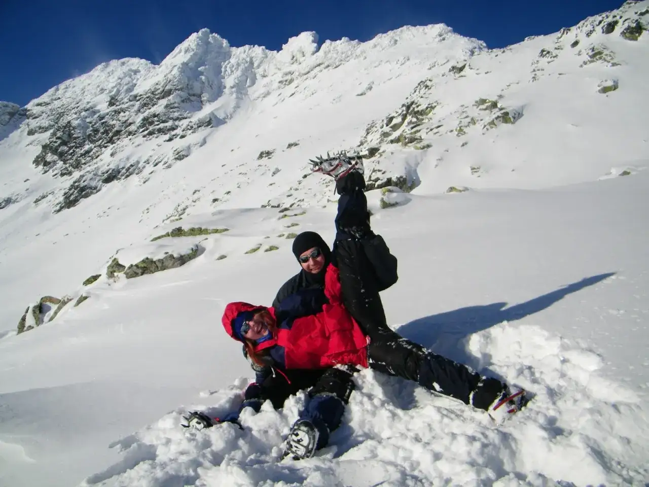 Szaleństwo w śniegu w Tatrach w maju. Dwoje ludzi w zimowym stroju, z rakami na nogach, wygłupia się na tle ośnieżonych szczyt&oacute;w.