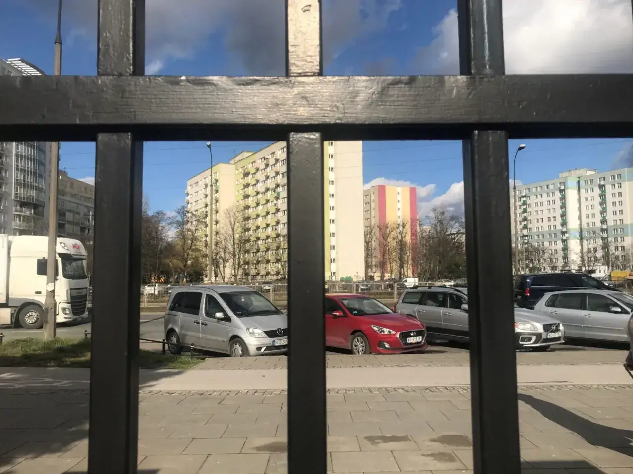 Widok przez kratę na parking we wspólnocie mieszkaniowej. Zaparkowane samochody i budynki mieszkalne.
