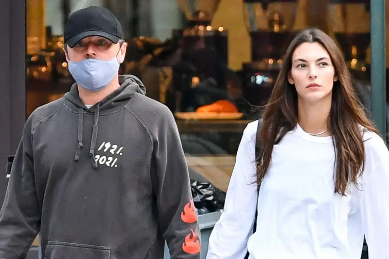 Leonardo DiCaprio w masce i czapce idzie z brunetką w białej bluzce.