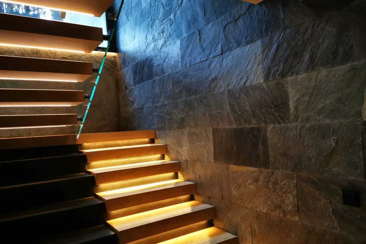 Nowoczesne schody z drewna i szkła, z podświetlanymi stopniami, przylegają do ściany wyłożonej kamieniem.