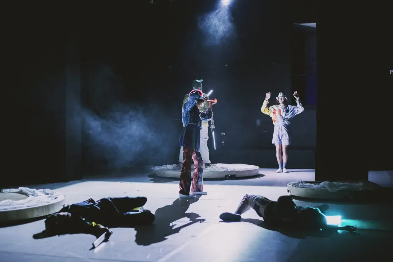 Scena teatralna z dymem i światłem. Na pierwszym planie leżą dwie postacie. W głębi stoi osoba z instrumentem, a obok niej druga z podniesionymi rękami. Sprawdź teatr muzyczny warszawa repertuar.